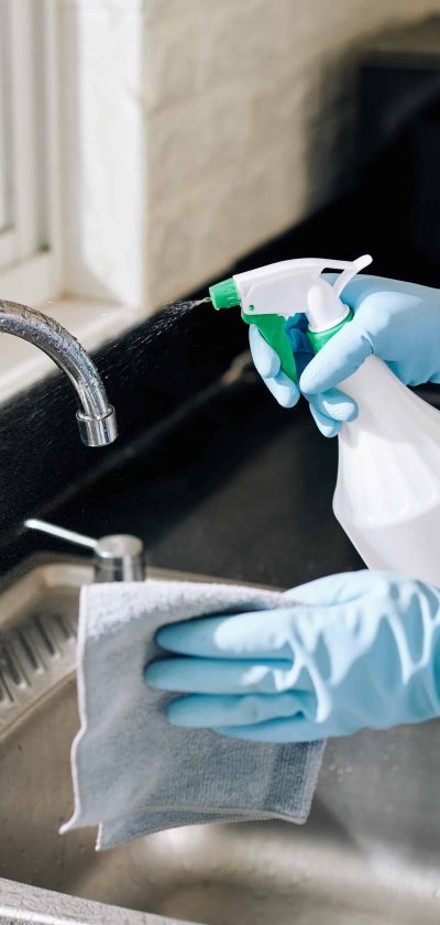 Hands of housewife spraying cleaning detergent on kitchen tap and wiping it with soft cloth