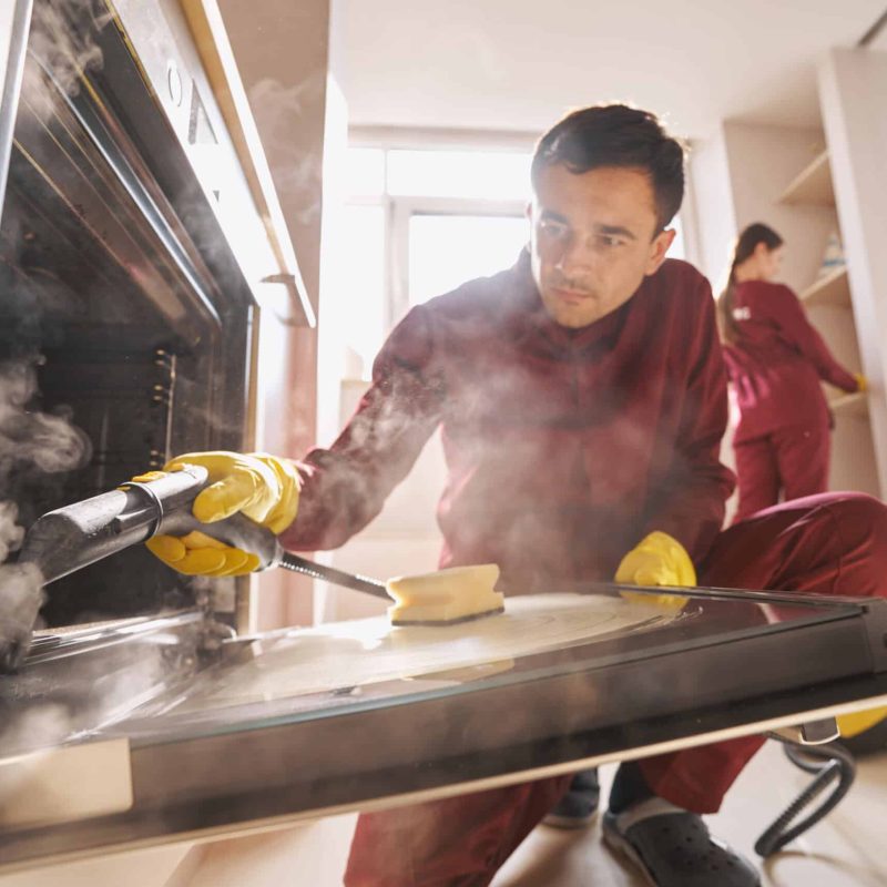 Cleaner using steam-cleaning device for disinfecting open oven door glass while his coworker dusting